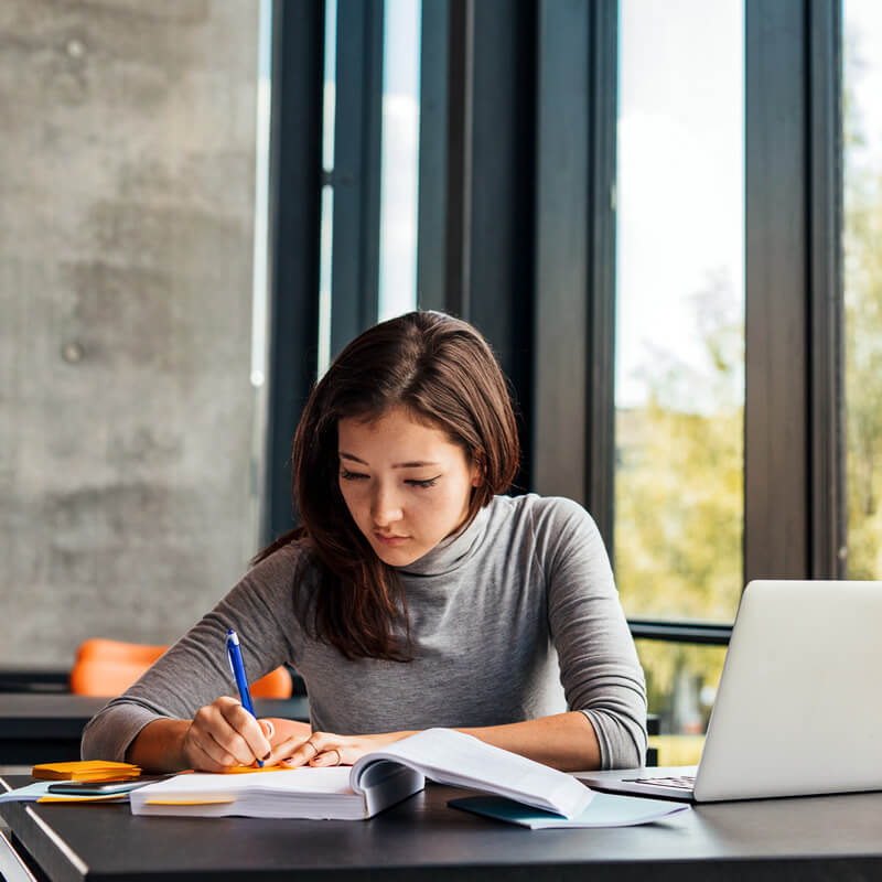 A college student writing notes at a desk in a library.