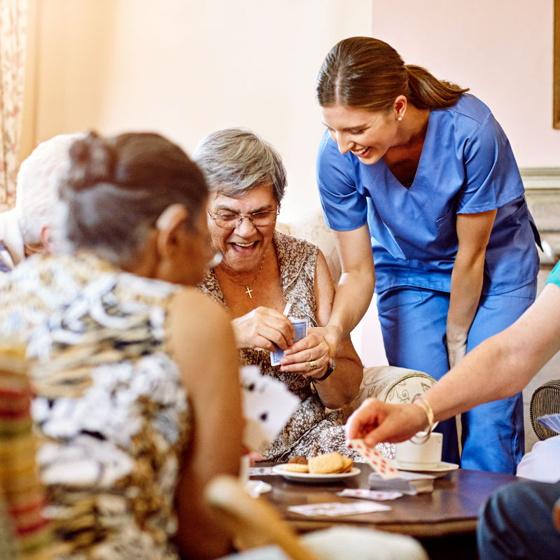 Older women playing cards, having tea with healthcare provider nearby.