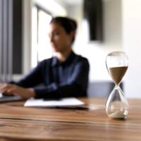 An hourglass sitting on a desk as a person works at a computer.